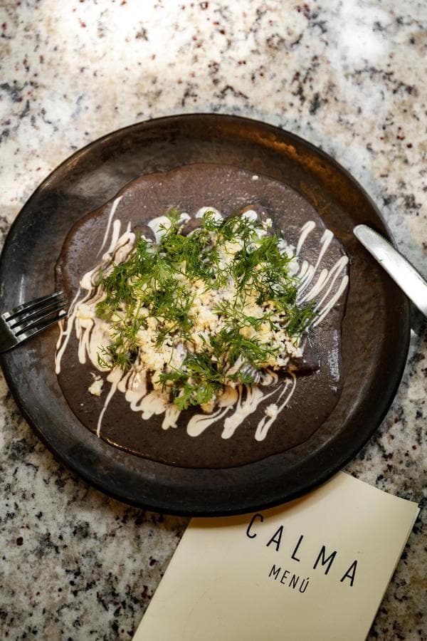 Un plato de barro oscuro con verduras y crema, junto a la carta de Calma